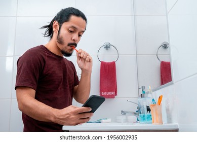 Young Latin Man With Long Black Hair Using The Phone While Brushing Teeth At The Bathroom