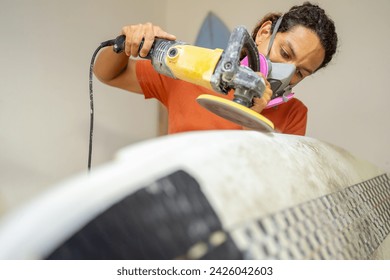 Young latin man with protective face mask grinding the edges of a surfboard in a shop - Powered by Shutterstock