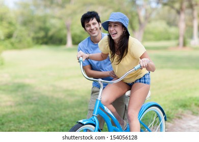 Young Latin Man In Shorts And Blue Shirt Helps Young Woman Riding Blue Bike With Green Trees And Grass. Horizontal Shallow Focused Composition.