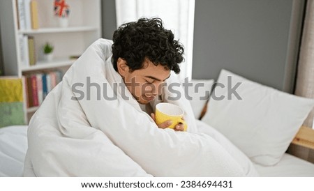 Young latin man smelling cup of coffee covering with blanket at bedroom
