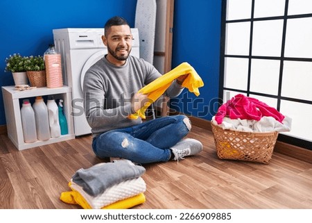 Young latin man smiling confident folding clothes at laundry room