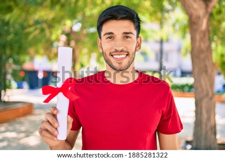 Young latin man smiling happy holding graduated diploma standing at the city.