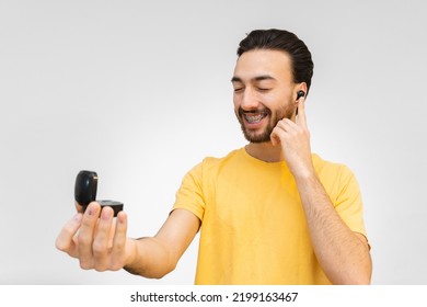 Young Latin Man Smiling With Wireless, Touch-sensitive Ear Buds Out Of Rechargeable Case. On White Background.