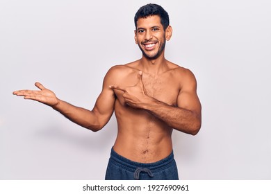 Young latin man standing shirtless amazed and smiling to the camera while presenting with hand and pointing with finger. 