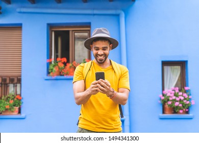Young Latin Man Using Cellphone Smiling Wearing Yellow T-Shirt Outdoors. Student Guy Holding Mobile Phone Over a Blue Background. Handsome Man Smiling. Lifestyle Concept.