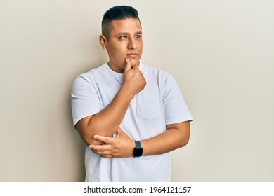 Young latin man wearing casual white t shirt with hand on chin thinking about question, pensive expression. smiling with thoughtful face. doubt concept. 