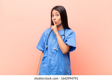 young latin nurse asking for silence and quiet, gesturing with finger in front of mouth, saying shh or keeping a secret against pink wall