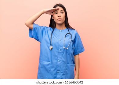 Young Latin Nurse Greeting The Camera With A Military Salute In An Act Of Honor And Patriotism, Showing Respect Against Pink Wall