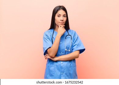 Young Latin Nurse Looking Serious, Confused, Uncertain And Thoughtful, Doubting Among Options Or Choices Against Pink Wall