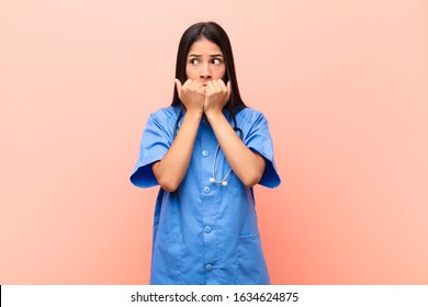 Young Latin Nurse Looking Worried, Anxious, Stressed And Afraid, Biting Fingernails And Looking To Lateral Copy Space Against Pink Wall