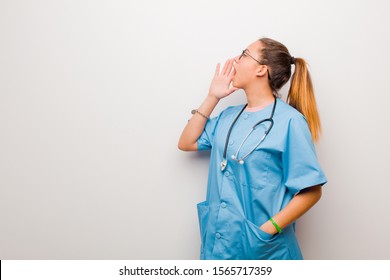 Young Latin Nurse Profile View, Looking Happy And Excited, Shouting And Calling To Copy Space On The Side Against White Wall