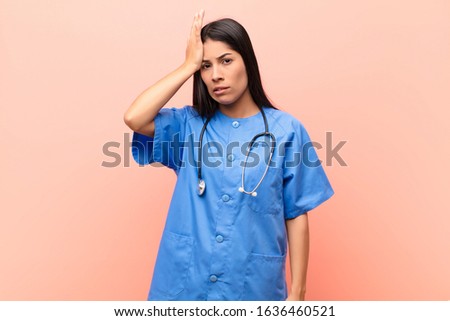 young latin nurse raising palm to forehead thinking oops, after making a stupid mistake or remembering, feeling dumb against pink wall