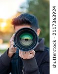 Young latin person pointing directly at you with his camera lens. Photographer taking pictures. Vertical shoot on natura. Person pointing a camera lens at you