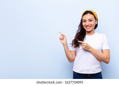 young latin pretty woman smiling happily and pointing to side and upwards with both hands showing object in copy space against flat wall