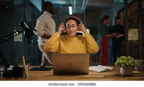 Young Latin Project Manager Putting On Headphones and Working on Laptop Computer in Busy Creative Office Environment. Beautiful Diverse Multiethnic Female Specialist is Writing Business Strategy.