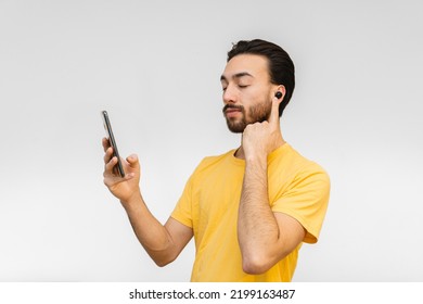 Young Latin Queer Man Connecting Wireless Touch Ear Buds To His Cell Phone. White Background