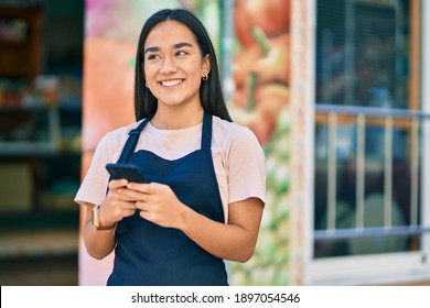 Young latin shopkeeper girl smiling happy using smartphone at fruit store.