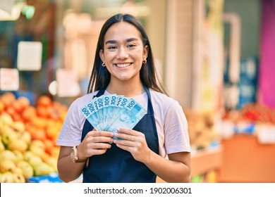 Young Latin Shopkeeper Girl Smiling Happy Holding Brazil Real Banknotes At The Fruit Store.