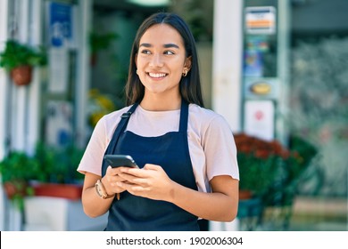 Young latin shopkeeper girl smiling happy using smartphone at florist.
