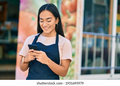 Young Latin Shopkeeper Girl Smiling Happy Using Smartphone At Fruit Store.