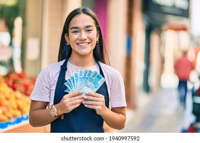 Young Latin Shopkeeper Girl Smiling Happy Holding Brazil Real Banknotes At The Fruit Store.