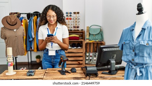 Young latin shopkeeper woman using smartphone working at clothing store.