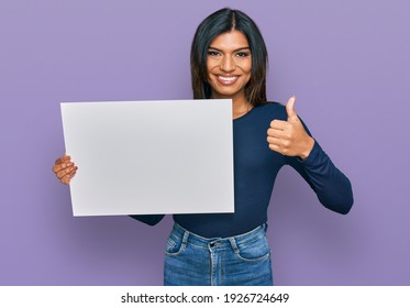Young latin transsexual transgender woman holding blank empty banner smiling happy and positive, thumb up doing excellent and approval sign 