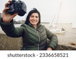 young latin traveler woman tourist happy smiling taking a selfie with her DSRL digital camera and with the river and boats in the background, travel and technology concept, copy space.