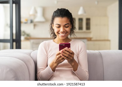 Young Latin Woman Chatting By The Phone And Smiling Widely, Sitting On A Comfortable Sofa At Home, Got Message From Family, Talking By Video, Long-distance Relationship, Light Kitchen Behind The Back