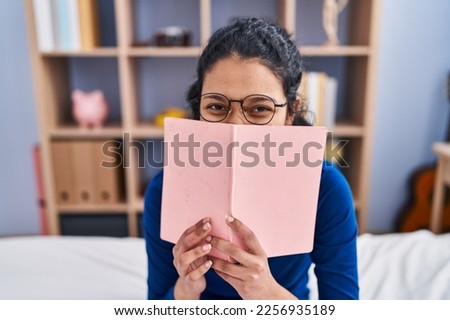 Young latin woman covering mouth with book sitting on bed at bedroom