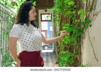 young Latin woman with curlers, elegant, is standing at entrance of house looking at and touching fresh leaves of climbing plant that is on wall in entrance hall - Powered by Shutterstock