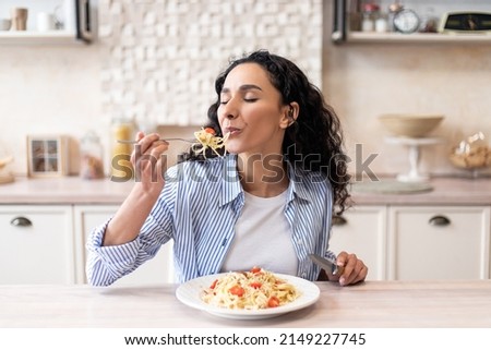 Young latin woman eating delicious pasta, enjoying tasty homemade lunch with closed eyes while sitting at table in light kitchen interior, free space