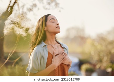 Young latin woman with hand on chest breathing in fresh air in a beautiful garden during sunset. Healthy mexican girl enjoying nature while meditating during morning exercise routine with closed eyes.