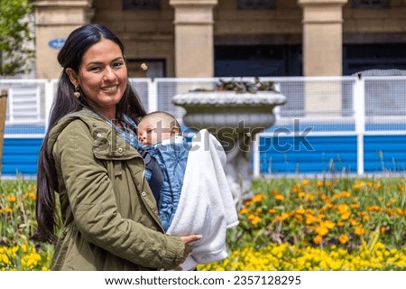 Young latin woman with her baby in a baby carrier, in a park with yellow flowers.