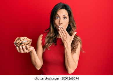 Young latin woman holding bowl with cookies covering mouth with hand, shocked and afraid for mistake. surprised expression 