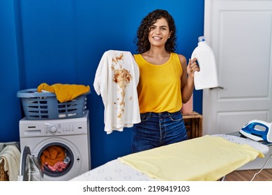 Young Latin Woman Holding Dirty T Shirt And Detergent Bottle At Laundry Room