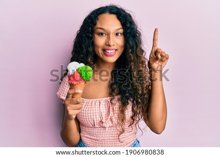 Young latin woman holding ice cream smiling with an idea or question pointing finger with happy face, number one 