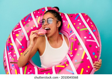 Young latin woman holding an inflatable donut eating a donut isolated on blue background