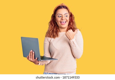 Young latin woman holding laptop pointing thumb up to the side smiling happy with open mouth  - Shutterstock ID 1871149333