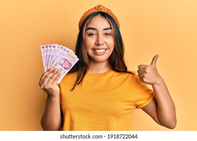 Young latin woman holding mexican pesos banknotes smiling happy and positive, thumb up doing excellent and approval sign 