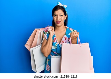 Young Latin Woman Holding Shopping Bags Clueless And Confused Expression. Doubt Concept. 