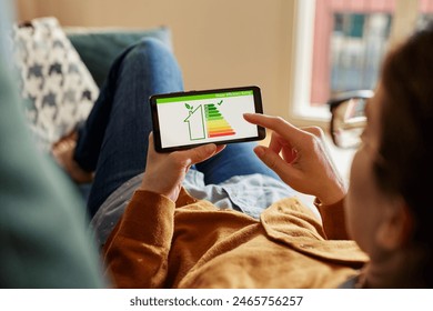 Young latin woman holding smartphone with energy efficiency chart and class on screen. Hispanic girl holding smart phone and looking at house efficiency rating. Bio energetic house concept. - Powered by Shutterstock