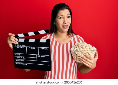 Young Latin Woman Holding Video Film Clapboard And Popcorn Clueless And Confused Expression. Doubt Concept. 