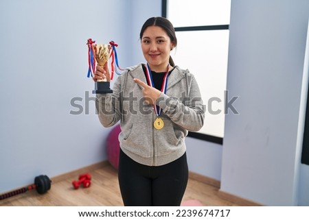 Young latin woman holding winner trophy smiling happy pointing with hand and finger 