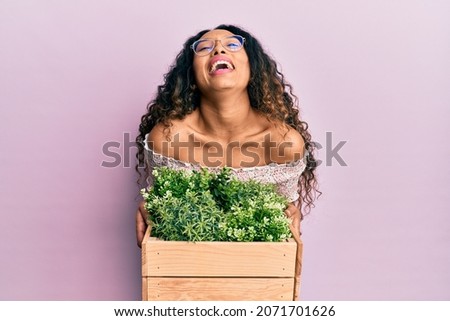 Young latin woman holding wooden plant pot smiling and laughing hard out loud because funny crazy joke. 
