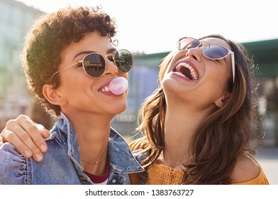 Young latin woman laughing while friend inflating bubble gum. Closeup face of multiethnic friends enjoying outdoor street. Brazilian girl laughing and blowing chewing gum with friend embracing her. - Shutterstock ID 1383763727