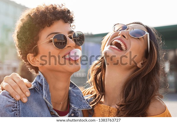 Young latin woman laughing while friend inflating\
bubble gum. Closeup face of multiethnic friends enjoying outdoor\
street. Brazilian girl laughing and blowing chewing gum with friend\
embracing her.