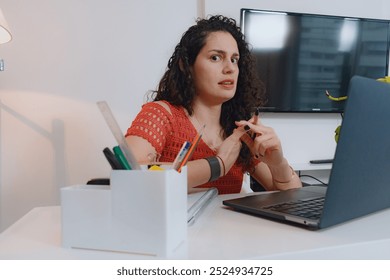 Young Latin woman in red and curly hair, works on the laptop, looks at the camera worried while doing accounts with her hands, looks with a face of doubt and disapproval, working in her home office - Powered by Shutterstock