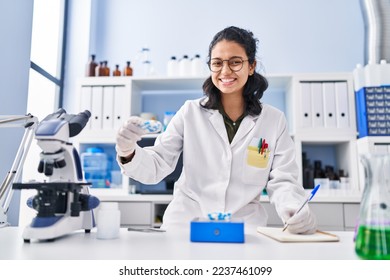 Young latin woman scientist writing on notebook holding pills at laboratory - Powered by Shutterstock