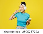 Young Latin woman she wears blue top t-shirt headband casual clothes listen to music in headphones use mobile cell phone sing song in microphone isolated on plain yellow background. Lifestyle concept